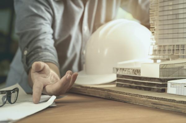 Image of a person pointing to a document and holding a white hard hat, leaning against a table